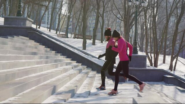 Couple of athletes running up on concrete staircase in city park in slow motion, snow lying on ground. Tracking shot joggers training together in winter. Concept of fitness