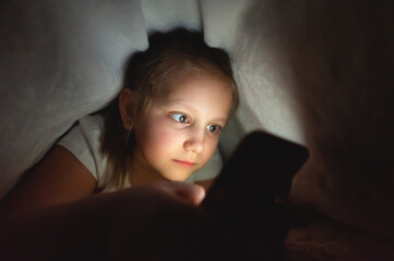 Child watching video smartphone under the blanket on the bed at night when light flashes bounce off the screen, children use addiction games and cartoon concept