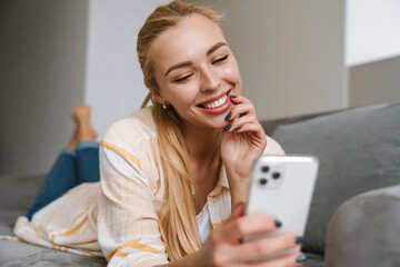 Happy blonde woman smiling and using cellphone while lying on couch