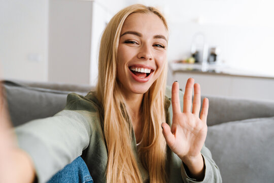 Happy blonde woman waving hand while taking selfie photo at home