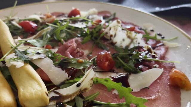 Close-up shot of enjoying Italian meal in the restaurant. Appetizing dish with prosciutto, parmesan, rocket salad and breadsticks aside