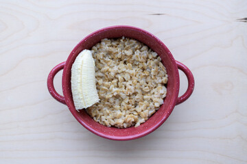 A plate of cooked oatmeal and a slice of banana. Concept of breakfast, diet, vegetarian food. Background - light wood, top view.