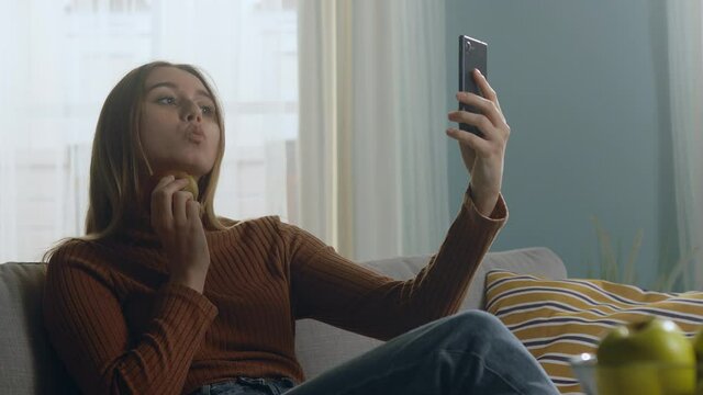 Young woman in blue jeans and brown sweater sits on beige sofa on window background, holding red apple and taking selfie. Average static plan.