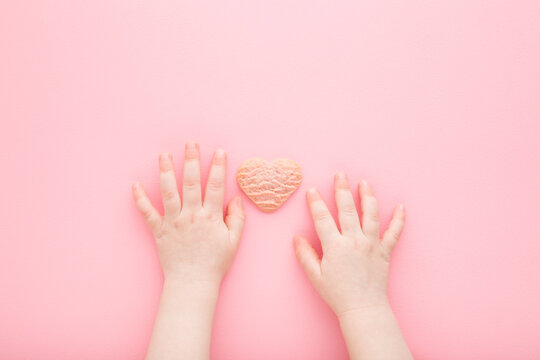 Baby Hands And Heart Shape Of Dry Cookie On Light Pink Table Background. Pastel Color. Sweet Snack. Closeup. Point Of View Shot. Top Down View.