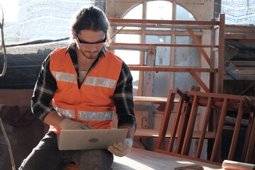 A young carpenter is looking at an order in a laptop computer to produce wooden furniture in his wood factory.