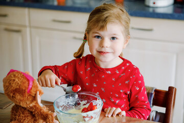 Cute little toddler girl eating fresh strawberries with cotton cheese or yogurt in domestic kitchen. Happy child tasting healthy seasonal berries and fruits, indoors.