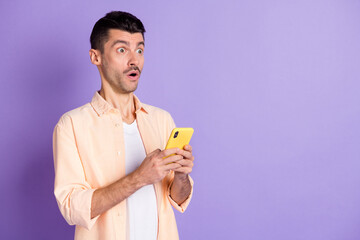 Photo of impressed brunet guy hold telephone look empty space wear peach shirt isolated on lilac color background