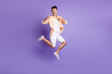 Fototapeta na wymiar Full length photo of man jump raise two thumbs up wear beige shirt shorts sneakers isolated purple color background