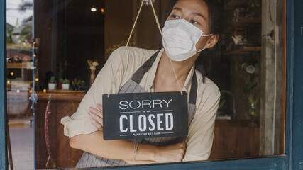 Young Asia girl wear face mask turning a sign from open to closed sign on glass door cafe after...