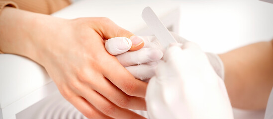 Obraz na płótnie Canvas Manicure master wears white gloves with nail file doing manicure on female fingernails in a nail salon