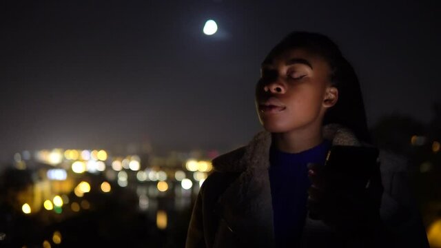 A young black woman works on a smartphone and thinks in an urban area at night - the moon and city lights in the blurry background
