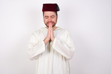 Indoor closeup of young handsome Caucasian man wearing Arab djellaba and Fez hat over white wall...