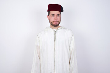 Amazed puzzled young handsome Caucasian man wearing Arab djellaba and Fez hat over white wall, curves lips and has worried look, sees something awful in front.