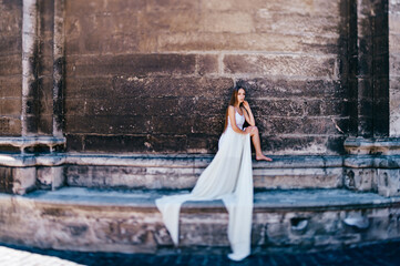 Young romantic elegant girl in long white flowy dress posing over stone ancient wall