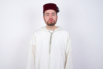 young handsome Caucasian man wearing Arab djellaba and Fez hat over white wall, keeps lips as going to kiss someone, has glad expression, grimace face. Standing indoors. Beauty concept.