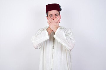 Upset young handsome Caucasian man wearing Arab djellaba and Fez hat over white wall, covering her mouth with both palms to prevent screaming sound, after seeing or hearing something bad.