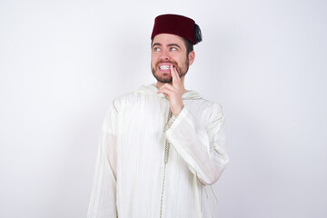 young handsome Caucasian man wearing Arab djellaba and Fez hat over white wall with thoughtful expression, looks to the camera, keeps hand near face, bitting a finger thinks about something pleasant.