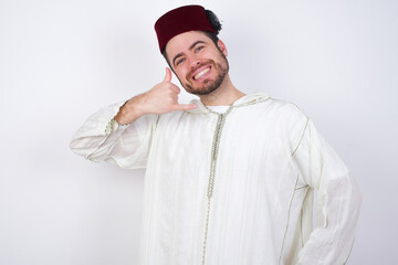 young handsome Caucasian man wearing Arab djellaba and Fez hat over white wall imitates telephone conversation, makes phone call gesture with hands, has confident expression. Call me!
