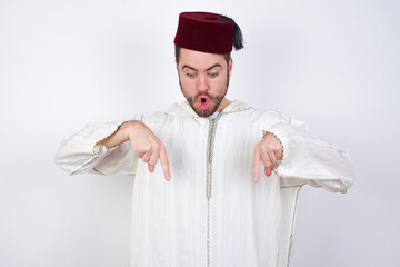 Amazed young handsome Caucasian man wearing Arab djellaba and Fez hat over white wall points down...