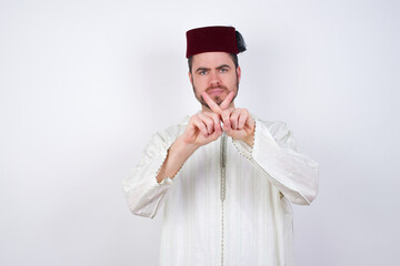 young handsome Caucasian man wearing Arab djellaba and Fez hat over white wall has rejection angry...