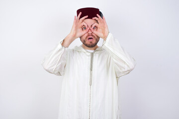 Playful excited young handsome Caucasian man wearing Arab djellaba and Fez hat over white wall showing Ok sign with both hands on eyes, pretending to wear spectacles.