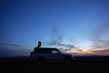 Fototapeta na wymiar Silhouette of girl with ponytail on roof of car on background of sunset sky. Blue and orange sky. White off road car