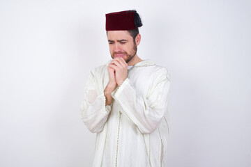 Sad young handsome Caucasian man wearing Arab djellaba and Fez hat over white wall feeling upset while spending time at home alone staring at camera with unhappy or regretful look.