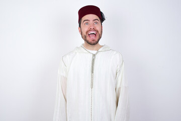 Surprised young handsome Caucasian man wearing Arab djellaba and Fez hat over white wall, shrugs shoulders, looking sideways, being happy and excited. Sudden reactions concept.