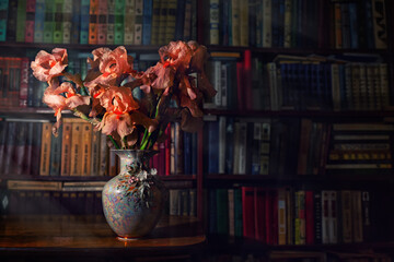 porcelain vase with pink irises stands on the table. There is a bookcase in the background. Vintage dark colors © _KUBE_