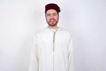 young handsome Caucasian man wearing Arab djellaba and Fez hat over white wall with happy and funny face smiling and showing tongue.