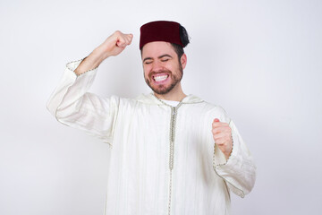 Attractive young handsome Caucasian man wearing Arab djellaba and Fez hat over white wall celebrating a victory punching the air with his fists and a beaming toothy smile.