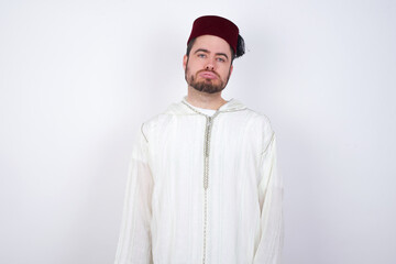Gloomy, bored young handsome Caucasian man wearing Arab djellaba and Fez hat over white wall frowns face looking up, being upset with so much talking hands down, feels tired and wants to leave.