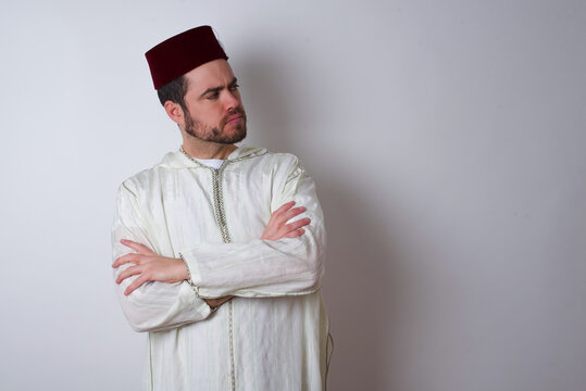 Image of upset young handsome Caucasian man wearing Arab djellaba and Fez hat over white wall with arms crossed. Looking with disappointed expression aside after listening to bad news.