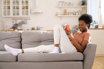 Happy Black woman enjoy lazy weekend, resting on cozy sofa at home, using smartphone. Young African...