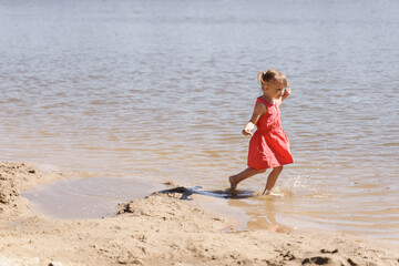 Fototapeta na wymiar little girl in a red dress runs along the seashore barefoot on a summer sunny day