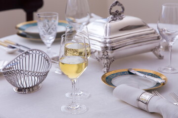 Glasses half filled with white wine on elegant table set with beautiful antique silver and porcelain dishes in soft focus background