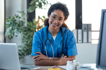 Attractive afro female doctor smiling looking at camera while working with laptop in the...