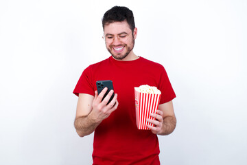 Smiling Young handsome man in red T-shirt against white background eating p using cell phone, messaging, being happy to text with friends, looking at smartphone. Modern technologies and communication.