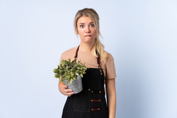 Blonde gardener woman holding a plant over isolated background having doubts and with confuse face expression