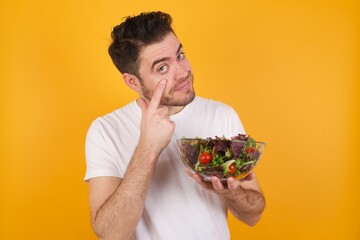 young handsome Caucasian man holding a salad bowl against yellow wall, looking, observing, keeping an eye on an object in front, or watching out for something.