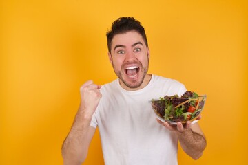 young handsome Caucasian man holding a salad bowl against yellow wall rejoicing success and victory clenching his fists with joy being happy to achieve her aim and goals. Positive emotions, feelings.