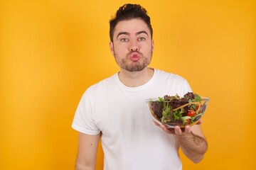 young handsome Caucasian man holding a salad bowl against yellow wall, keeps lips as going to kiss someone, has glad expression, grimace face. Standing indoors. Beauty concept.