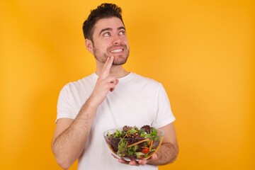 young handsome Caucasian man holding a salad bowl against yellow wall with thoughtful expression, looks to the camera, keeps hand near face, bitting a finger thinks about something pleasant.
