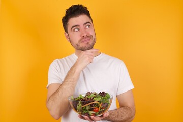 Dreamy young handsome Caucasian man holding a salad bowl against yellow wall with pleasant expression, looks sideways, keeps hand under chin, thinks about something pleasant.