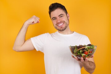 Smiling young handsome Caucasian man holding a salad bowl against yellow wall raises hand to show muscles, feels confident in victory, strong and independent.