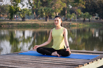 Young Asian beautiful woman practicing yoga and meditate in the lotus pose outdoor beside the lake in the morning for relaxation and peace of mind. Harmony and meditation concept. Healthy lifestyle