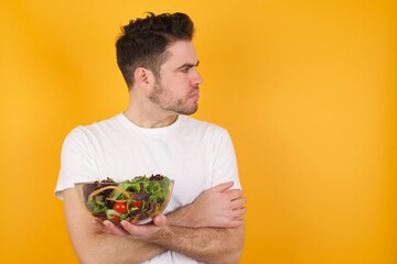 Image of upset young handsome Caucasian man holding a salad bowl against yellow wall with arms crossed. Looking with disappointed expression aside after listening to bad news.