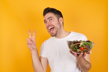 Isolated shot of cheerful young handsome Caucasian man holding a salad bowl against yellow wall makes peace or victory sign with both hands, feels cool.