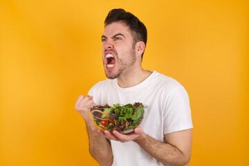 young handsome Caucasian man holding a salad bowl against yellow wall excited and glad to achieve victory, clenches fists, screams in excitement with closed eyes,successful person.