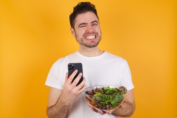 Pleased young handsome Caucasian man holding a salad bowl against yellow wall using self phone and looking and winking at the camera. Flirt and coquettish concept.